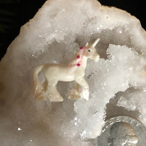 May include: A small, white unicorn figurine with a pink mane and glitter accents. The unicorn is set against a white, crystalline background, with a quarter coin for size comparison. The figurine has a horn and is in a walking pose.