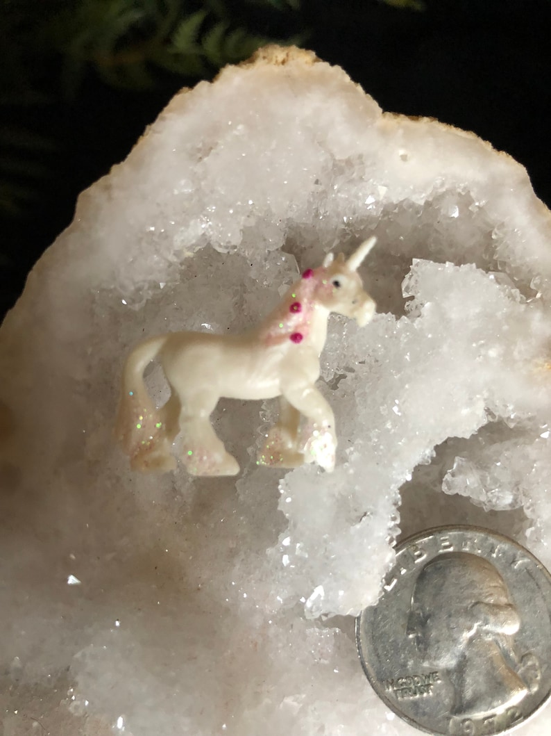 May include: A small, white unicorn figurine with a pink mane and glitter accents. The unicorn is set against a white, crystalline background, with a quarter coin for size comparison. The figurine has a horn and is in a walking pose.