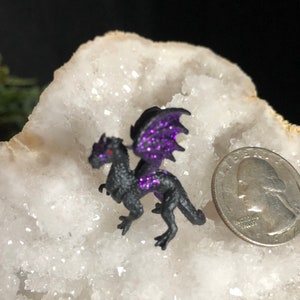May include: A small, black dragon figurine with purple glitter wings and accents. The dragon has red eyes and is posed on a white, crystalline surface. A U.S. quarter coin is next to the dragon for size comparison.