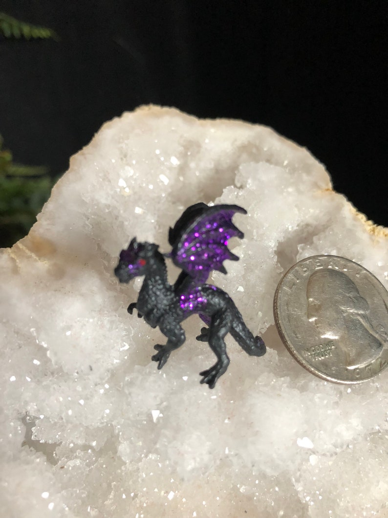 May include: A small, black dragon figurine with purple glitter wings and accents. The dragon has red eyes and is posed on a white, crystalline surface. A U.S. quarter coin is next to the dragon for size comparison.