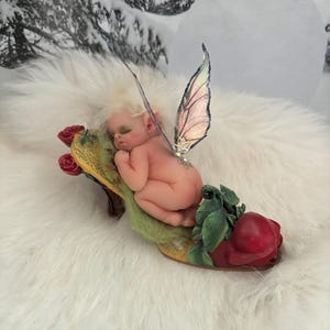 May include: A small, sleeping fairy doll with wings, lying on a red rose shoe. The fairy is nude and has a white wig. The shoe is yellow with green leaves and a red rose.