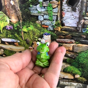 May include: A small green frog figurine wearing a crown, sitting with its hands clasped in front of it. The frog is sitting in a person's hand.
