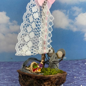 May include: A miniature sailboat made from a walnut shell with a white lace sail, a pink bow, and a gray felt mouse figure. The boat is sitting on a blue and white patterned surface.