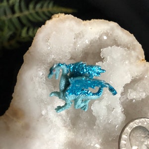 May include: A small, light blue dragon figurine with glittery wings and red eyes rests on a white, crystalline surface. The dragon is next to a US quarter coin. The background is dark with a hint of green foliage.