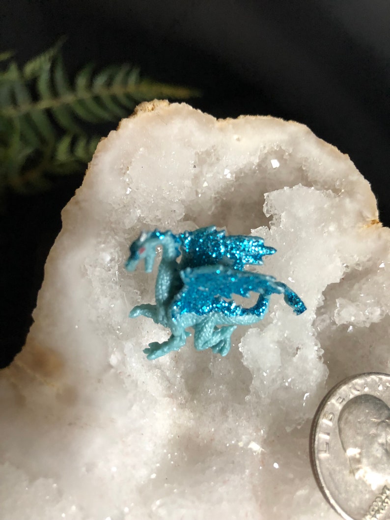 May include: A small, light blue dragon figurine with glittery wings and red eyes rests on a white, crystalline surface. The dragon is next to a US quarter coin. The background is dark with a hint of green foliage.