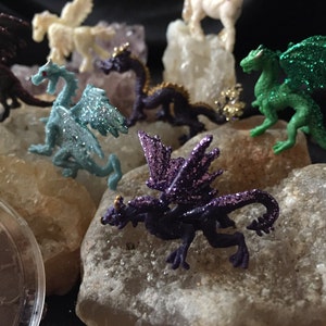 May include: A collection of miniature fantasy creatures, including dragons and pegasus figurines, in various colors like purple, green, and blue, with glitter accents. The figurines are displayed on light-colored rocks against a dark background.