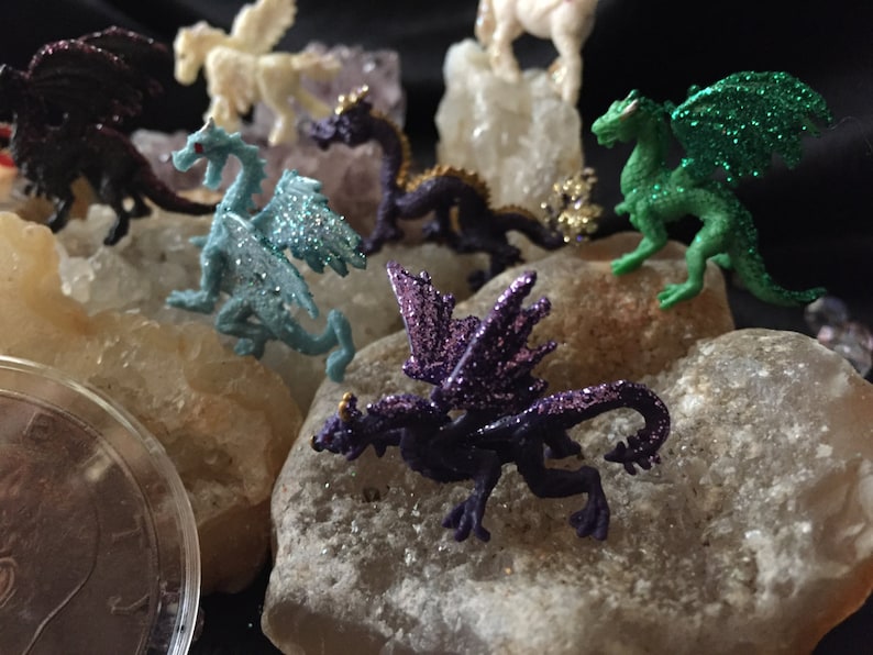 May include: A collection of miniature fantasy creatures, including dragons and pegasus figurines, in various colors like purple, green, and blue, with glitter accents. The figurines are displayed on light-colored rocks against a dark background.