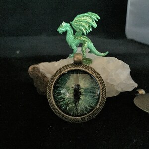 May include: A round pendant with a green eye design, set in a bronze-colored metal frame. A small green dragon figurine with glittery wings sits on a white stone above the pendant. The background is dark.