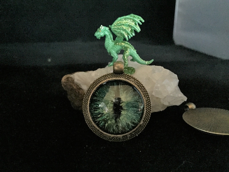 May include: A round pendant with a green eye design, set in a bronze-colored metal frame. A small green dragon figurine with glittery wings sits on a white stone above the pendant. The background is dark.