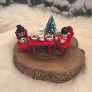 May include: A miniature Christmas scene featuring a snowman and a toy soldier seated at a red table. The table is set with small bowls and a decorative tree. The scene is set on a wooden slice, with a snowy background.