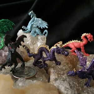May include: A collection of small, colorful dragon figurines. The dragons are in various colors, including green, blue, red, and purple, with glitter accents. They are posed on a bed of white and tan rocks, with a black background.