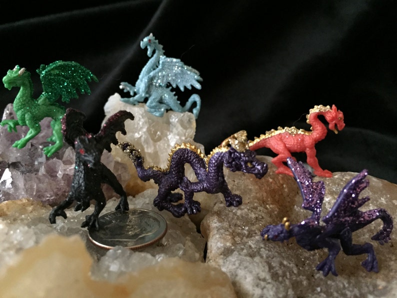 May include: A collection of small, colorful dragon figurines. The dragons are in various colors, including green, blue, red, and purple, with glitter accents. They are posed on a bed of white and tan rocks, with a black background.