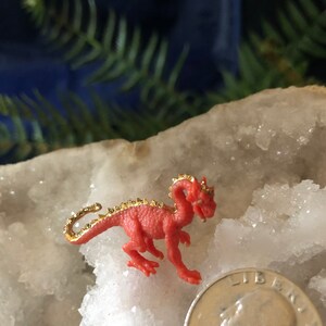 May include: A small, red dragon figurine with gold accents on its back and tail. The dragon is posed on a white, crystalline surface, next to a U.S. quarter coin for size comparison. The background is a dark blue.