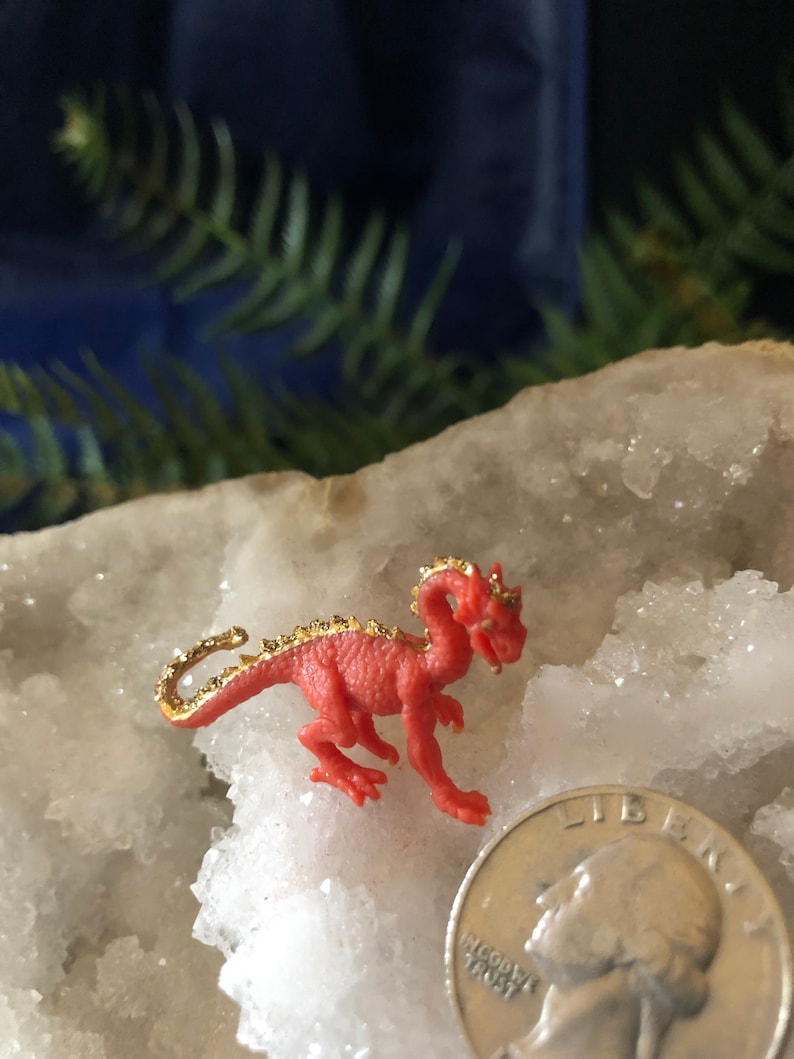 May include: A small, red dragon figurine with gold accents on its back and tail. The dragon is posed on a white, crystalline surface, next to a U.S. quarter coin for size comparison. The background is a dark blue.