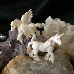 May include: Two small, white unicorn figurines with glitter accents and pink details are displayed on a bed of crystals. One unicorn has wings. The crystals are various shades of purple, white, and tan, creating a whimsical scene.