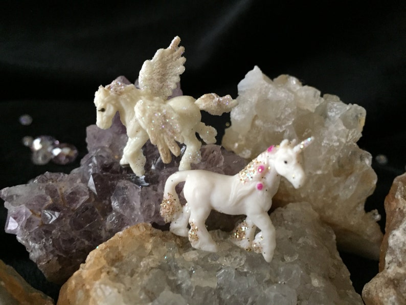 May include: Two small, white unicorn figurines with glitter accents and pink details are displayed on a bed of crystals. One unicorn has wings. The crystals are various shades of purple, white, and tan, creating a whimsical scene.