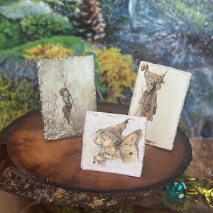 May include: Three small, framed fairy art prints. The prints feature whimsical illustrations of fairies and figures with delicate details. The frames have glittery edges. The prints are displayed on a wooden slice.