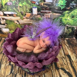 May include: A sleeping fairy figurine with purple hair and feathered accents rests in a deep purple flower. The fairy has delicate wings and is nestled in a detailed, handcrafted floral sculpture.