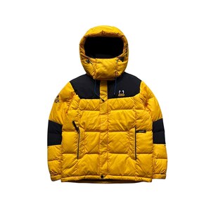 May include: A yellow puffer jacket with a black hood and shoulder panels. The jacket features a hood, a front zip, and multiple pockets. A logo is visible on the chest and left sleeve. The jacket is designed for cold weather conditions.