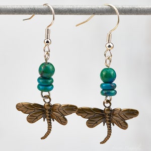 Op de afbeelding: Een paar oorhangers met een bronzen libellenbedel en turquoise kralen. De oorbellen zijn zilverkleurig en hebben een haaksluiting.
