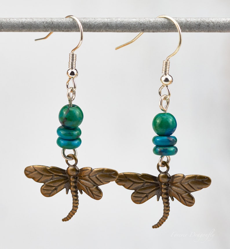 Op de afbeelding: Een paar oorhangers met een bronzen libellenbedel en turquoise kralen. De oorbellen zijn zilverkleurig en hebben een haaksluiting.