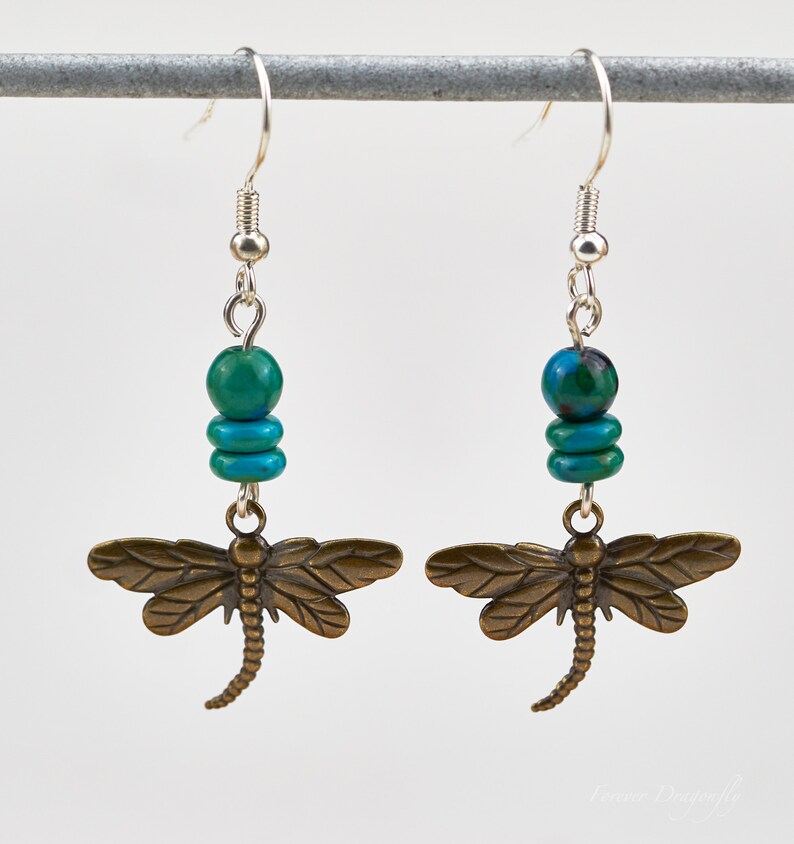 Op de afbeelding: Een paar oorhangers met een bronzen libellenbedel en turquoise kralen. De oorbellen hebben zilveren haakjes.