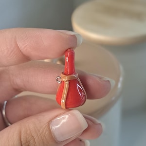 May include: A small, red bottle-shaped ornament with a tan band and a silver loop. The bottle has a rounded base and a long neck. It appears to be handmade, possibly a decorative item or a charm, and is held in a hand.