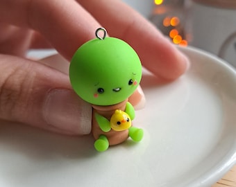 May include: A handmade polymer clay charm featuring a green character with a smiling face, holding a small yellow chick. The charm has a metal loop for hanging. The figure is sitting on a white plate.