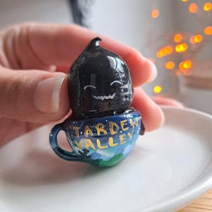 Puede incluir: Pequeña figura pintada a mano de un personaje negro sonriente sobre una taza azul. La taza está decorada con las palabras "Stardew Valley" en letras doradas, sobre un paisaje azul y verde.