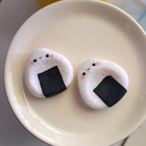 Könnte beinhalten: Zwei weiße, lächelnde Onigiri, oder Reisbällchen, mit schwarzen Algenakzenten und rosigen Wangen, liegen auf einem cremefarbenen Teller. Die Onigiri haben ein niedliches, cartoonartiges Design. Im Hintergrund befindet sich eine gelbe Vase mit Blumen.