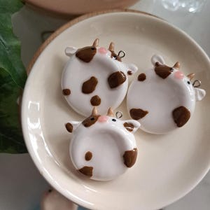 May include: Three white cow-shaped ornaments with brown spots, horns, and pink noses are arranged on a cream-colored plate. Each ornament has a small silver loop at the top. The ornaments are likely handmade and decorative.