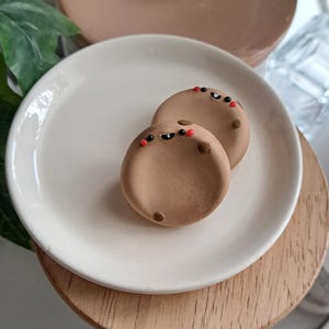 May include: Two brown, round ceramic pieces with black and red details, resembling small faces, sit on a white plate. The plate rests on a wooden stand, with a light brown lid and a beige plate in the background. The items appear to be decorative or for serving.