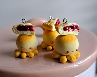 May include: Three miniature cherry pie octopus charms made of polymer clay. Each octopus has a yellow body with eight tentacles and a cherry pie on top. The pies are red and have a flaky crust.