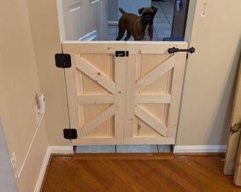 Può includere: Un cancello in legno con un design a X diagonale, dipinto in un colore legno chiaro. Il cancello ha cerniere nere e un chiavistello nero. Il cancello è installato in una porta, e un cane è visibile sullo sfondo.
