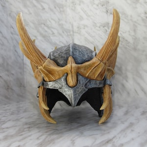 May include: A grey and brown helmet with horns. The helmet is made of a textured material and has a pointed top. The horns are long and curved.