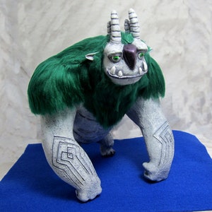 May include: A white and gray monster with green fur, two horns, and a black nose. The monster has green eyes and a toothy grin. The monster is standing on a blue surface.