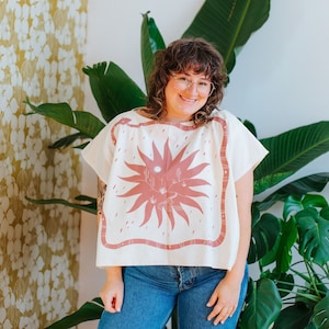 May include: A woman wearing a white shirt with a pink sun design and blue jeans. The shirt has a square neckline and short sleeves. The sun design is centered on the shirt and features a stylized sun with rays and a small moon in the center. The woman is smiling and looking at the camera.