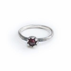 Peut inclure: Une bague en argent avec une pierre précieuse rouge foncé. La bague a un anneau simple et un sertissage à lunette qui maintient la pierre ronde et facettée. La bague est posée sur un fond blanc.