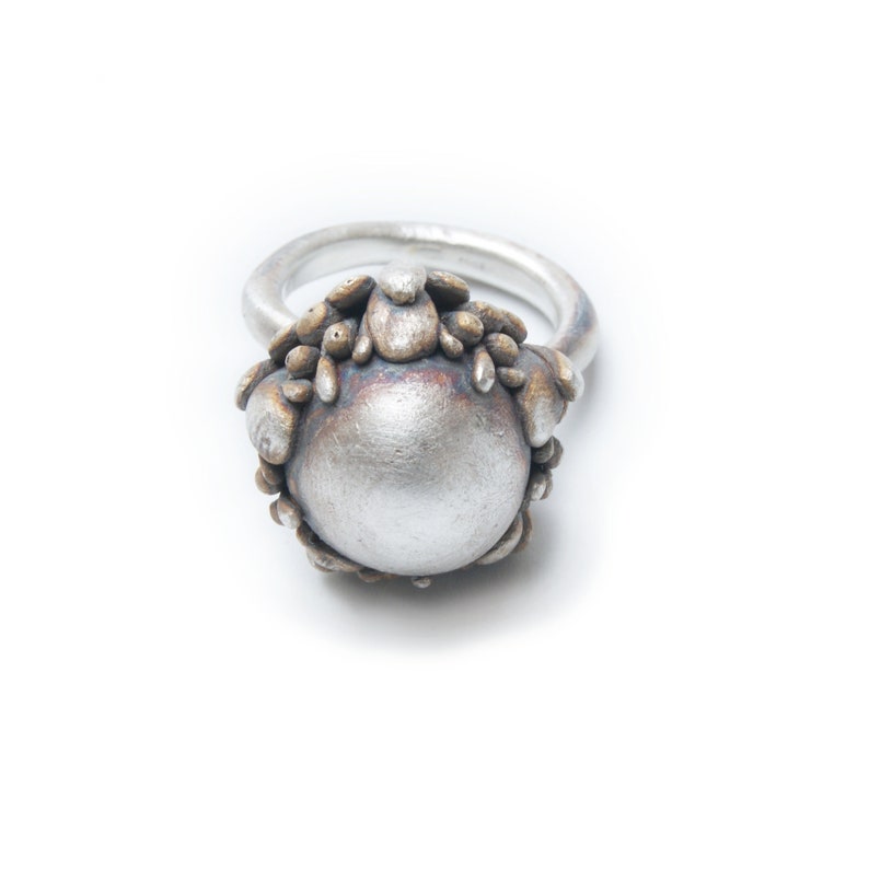 May include: A silver ring with a large, round, silver-colored stone surrounded by a textured, brown metal design.