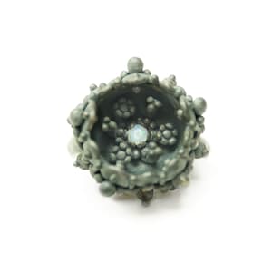 Op de afbeelding: Een handgemaakte ring met een getextureerd, donkergrijs metalen ontwerp. De ring heeft een centrale, lichtgekleurde opaalsteen, omringd door een gedetailleerde, organisch ogende structuur. De ring staat tegen een schone witte achtergrond.