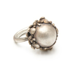 May include: A silver ring with a large, round, textured center piece. The center piece is surrounded by smaller, textured pieces that resemble pebbles.