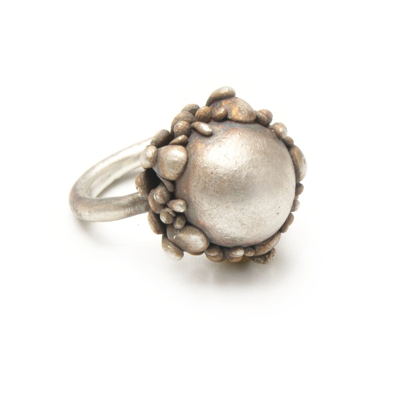 May include: A silver ring with a large, round, textured center piece. The center piece is surrounded by smaller, textured pieces that resemble pebbles.