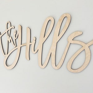 May include: Wooden wall art spelling "the Hills" in a cursive font. The letters are cut from light-colored wood and mounted on a white wall. The piece is designed for home decor.