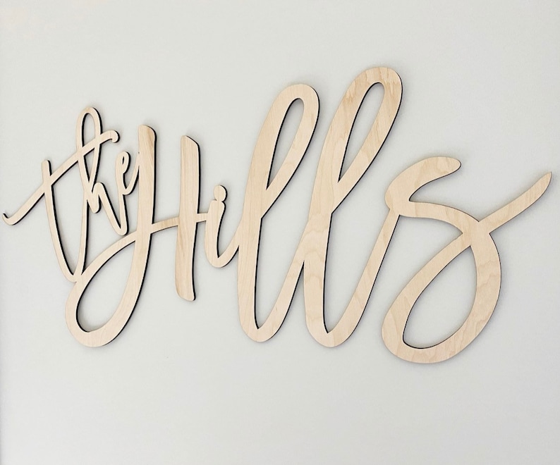 May include: Wooden wall art spelling "the Hills" in a cursive font. The letters are cut from light-colored wood and mounted on a white wall. The piece is designed for home decor.