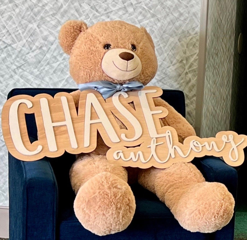 May include: A large, plush teddy bear with a light blue bow tie, seated in a dark blue armchair. A wooden sign with the name "CHASE" and "anthony" in a layered design is in front of the bear.
