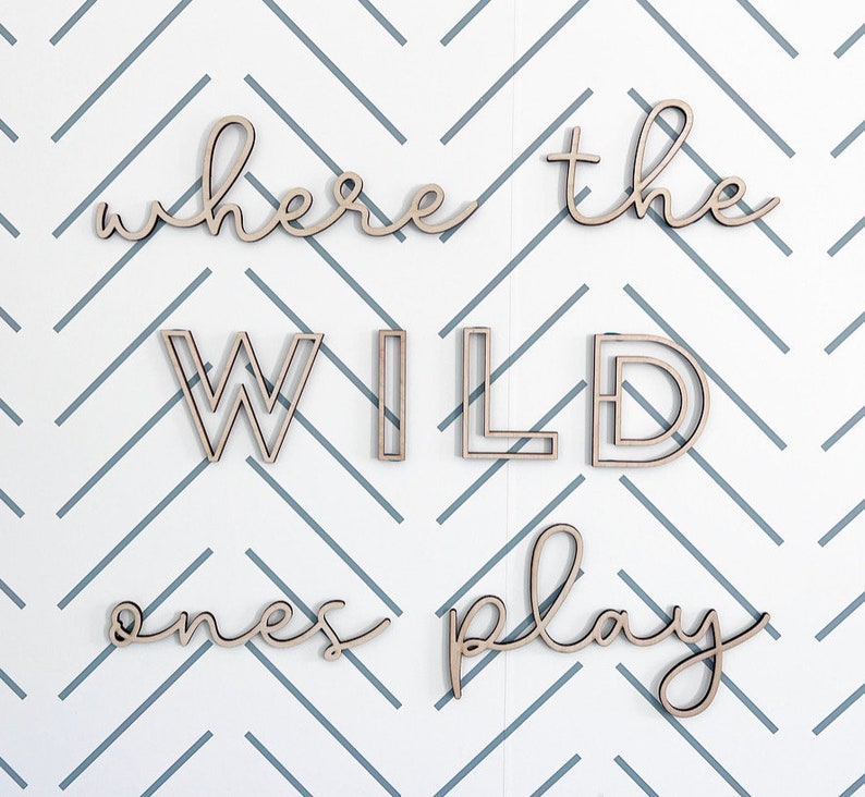 May include: Wooden wall art spelling out "where the wild ones play" in a cursive and block letter font. The letters are light brown and mounted on a white wall with a blue and gray chevron pattern.