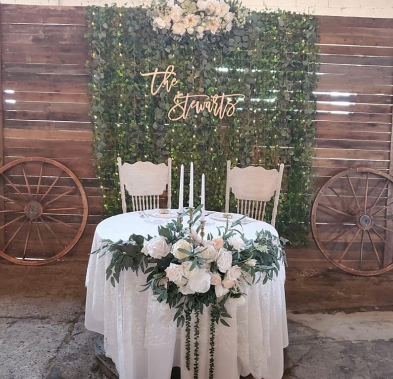 Pu&ograve; includere: Allestimento per ricevimento di nozze con un tavolo con una tovaglia di pizzo bianco, adornato con rose bianche e verde. Due sedie bianche di fronte al tavolo, su uno sfondo di verde e assi di legno. Il cartello recita "The Stewarts".