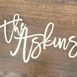 May include: Wooden sign with the words "the Askins" in a cursive font. The sign is light-colored and set against a dark wood grain background. The letters are connected, creating a flowing design.