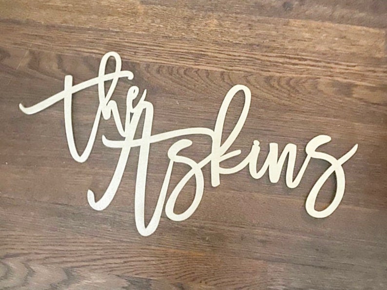 May include: Wooden sign with the words "the Askins" in a cursive font. The sign is light-colored and set against a dark wood grain background. The letters are connected, creating a flowing design.