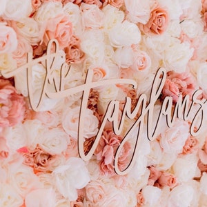 May include: A wooden sign with the name "Taylans" in cursive script is displayed against a backdrop of artificial flowers. The flowers are in shades of white, pink, and peach, creating a soft, romantic aesthetic.